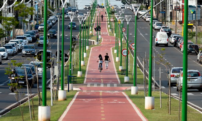 Avenida com alguns carros, no canteiro central ciclobia na cor vermelha e postes de iluminação na cor verde