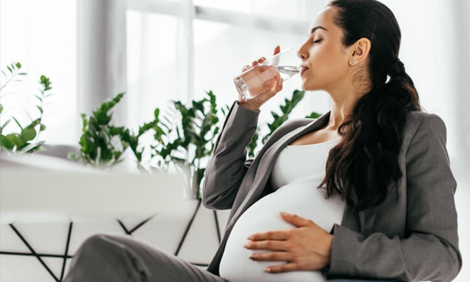 vista lateral de uma gestante tomando água sentada no sofá da sala de estar